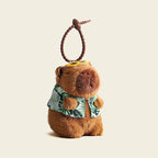 A plush capybara bag charm wearing a green and white Hawaiian shirt with leaf patterns, attached to a brown carabiner clip. The charm has a smiling face and is designed as a cute and trendy accessory for bags or backpacks.