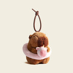 A plush capybara bag charm holding a pink swim ring with a small pig design, attached to a brown carabiner clip. This adorable accessory is perfect for adding a playful touch to bags or keychains.