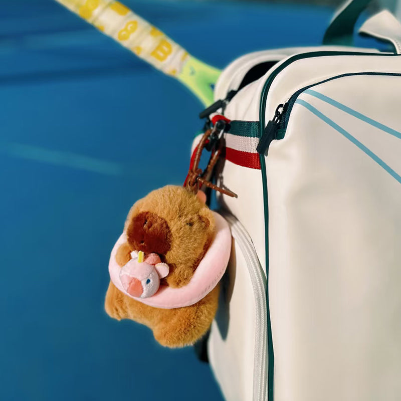 A cute brown capybara plush toy wearing a pink swimming ring and holding a small pink unicorn, hanging as a keychain on a white sports bag, with a tennis racket and blue court background visible.