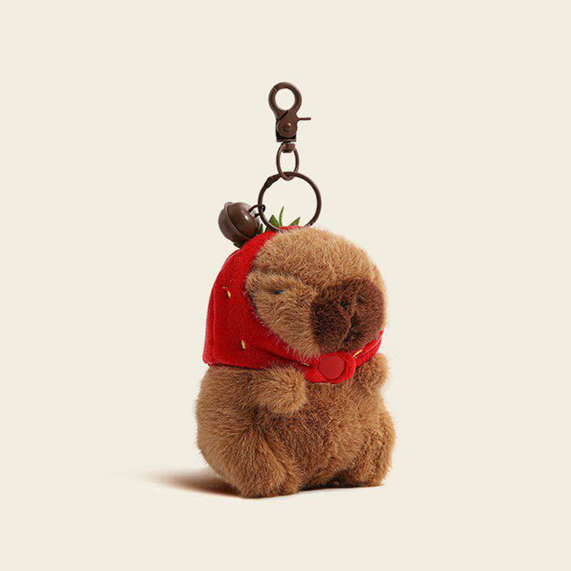 A plush capybara bag charm wearing a red strawberry hat with green leaves, attached to a brown carabiner clip. The charm has a smiling face and is dressed in a red outfit, making it a charming and whimsical addition to any bag or keychain.