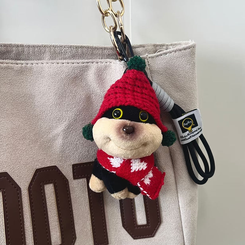 A plush dog bag charm wearing a red knitted hat with green pompoms and a matching red scarf with white patterns, attached to a black carabiner clip. This cute and festive accessory is designed to add a playful touch to bags or backpacks.