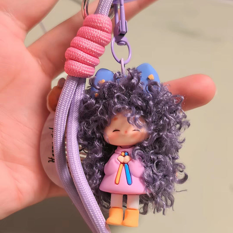 A close-up handheld shot of the purple curly-haired doll keychain. The image highlights the texture of the synthetic hair, the pink clothing details, and the thick lavender rope attachment.