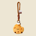 A cute, round, yellow plush cookie keychain with brown chocolate chips and a smiling face. It is attached to a brown braided rope with a metal ring and a small white tag featuring a logo.