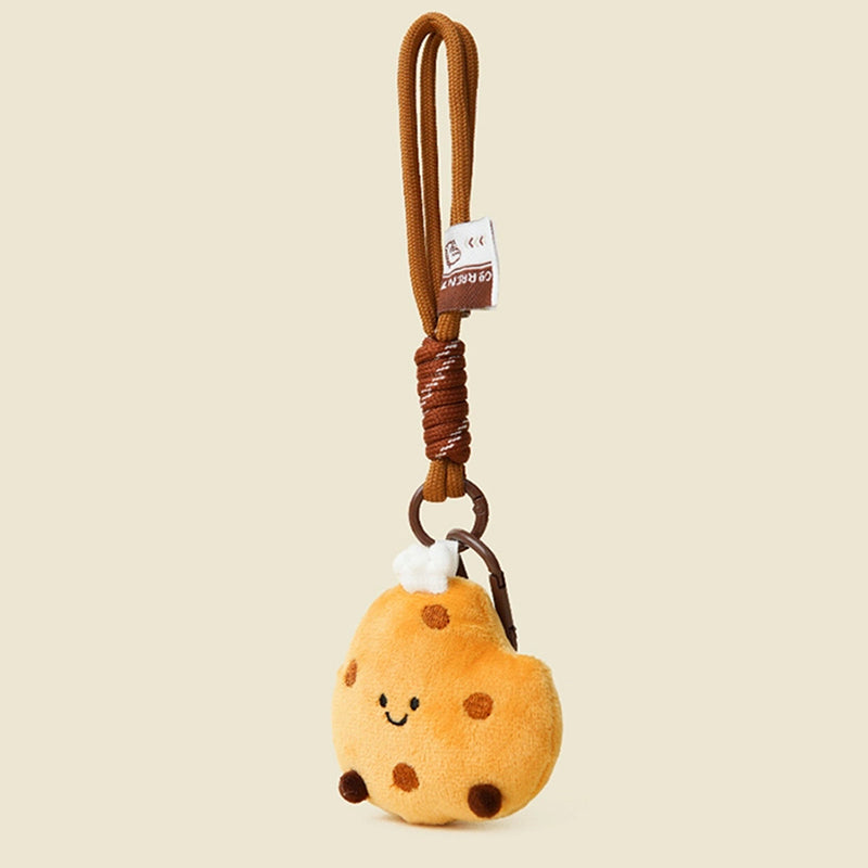A side view of a cute, round, yellow plush cookie keychain with brown chocolate chips and a smiling face. It is attached to a brown braided rope with a metal ring and a small white tag featuring a logo.