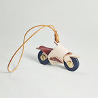 A side profile shot of a miniature leather motorcycle pendant. It has a cream body, maroon seat, and dark wheels, resting on a white surface with its long leather loop extending to the left.