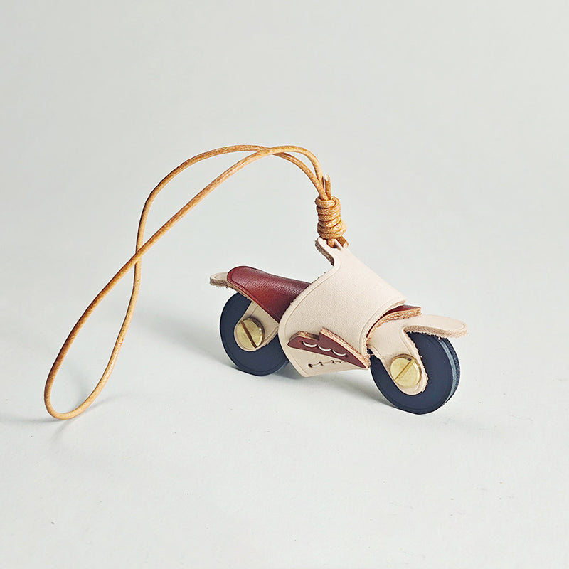A side profile shot of a miniature leather motorcycle pendant. It has a cream body, maroon seat, and dark wheels, resting on a white surface with its long leather loop extending to the left.