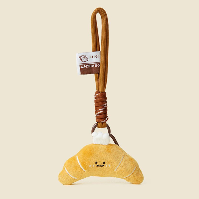 A cute, yellow plush croissant keychain with white stitched details and a smiling face. It is attached to a brown braided rope with a metal ring and a small white tag featuring a logo.