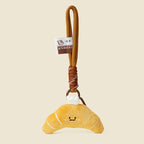 A cute, yellow plush croissant keychain with white stitched details and a smiling face. It is attached to a brown braided rope with a metal ring and a small white tag featuring a logo.