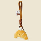 A side view of a cute, yellow plush croissant keychain with white stitched details and a smiling face. It is attached to a brown braided rope with a metal ring and a small white tag featuring a logo.