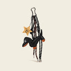 A black leather dachshund bag charm with orange accents on its paws and snout, attached to a silver cloud-shaped carabiner. It features two woven ropes—one white with black stripes, one black with white stripes—tied in knots, and a wooden star-shaped tag engraved with "You're a Star".