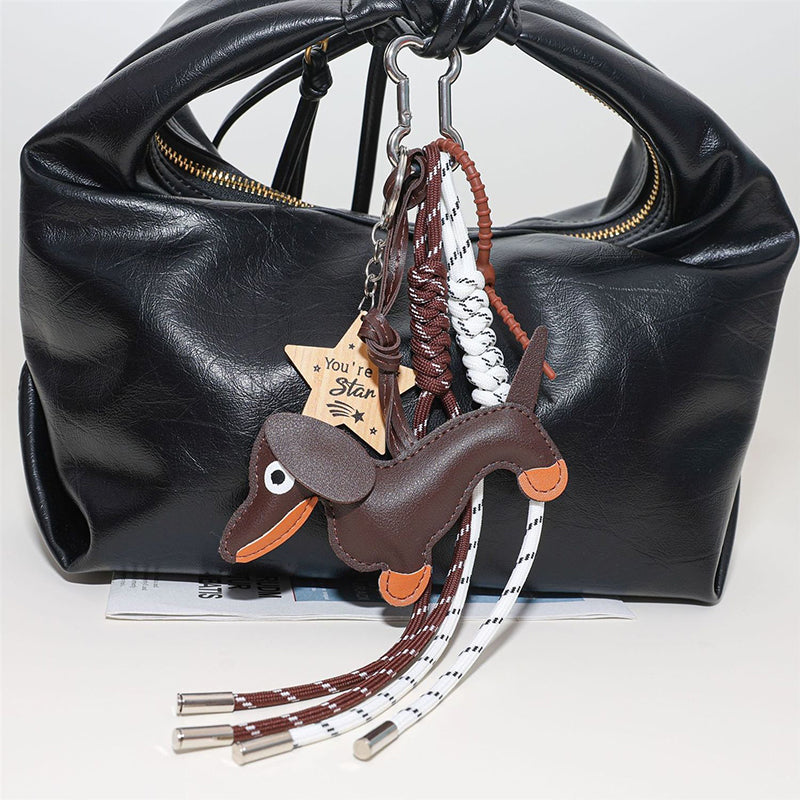 A brown leather dachshund bag charm with orange accents, hanging from the handle of a black leather tote bag. The charm is attached via a silver cloud-shaped carabiner and includes two woven ropes and a wooden star tag engraved with "You're a Star".