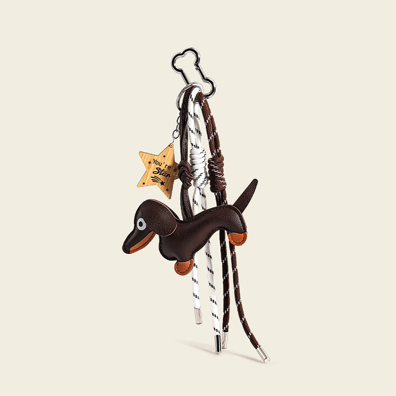 A brown leather dachshund bag charm with orange accents on its paws and snout, attached to a silver cloud-shaped carabiner. It features two woven ropes—one white with black stripes, one brown with white stripes—tied in knots, and a wooden star-shaped tag engraved with "You're a Star".