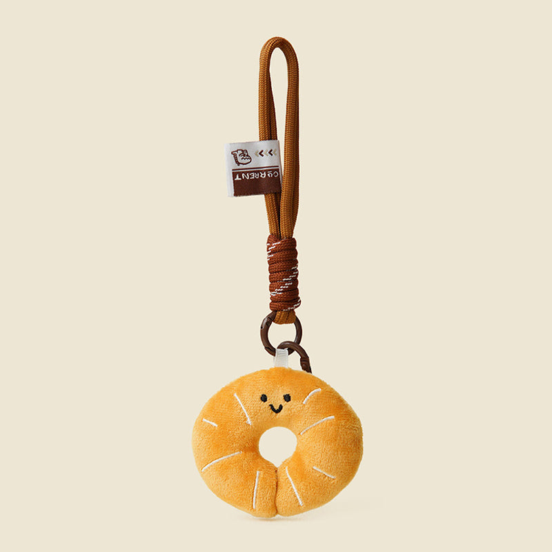 A cute, round, orange plush donut keychain with a smiling face and white stitched details. It is attached to a brown braided rope with a metal ring and a small white tag featuring a logo.