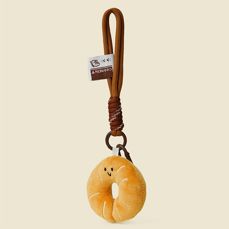 A side view of a cute, round, orange plush donut keychain with a smiling face and white stitched details. It is attached to a brown braided rope with a metal ring and a small white tag featuring a logo.