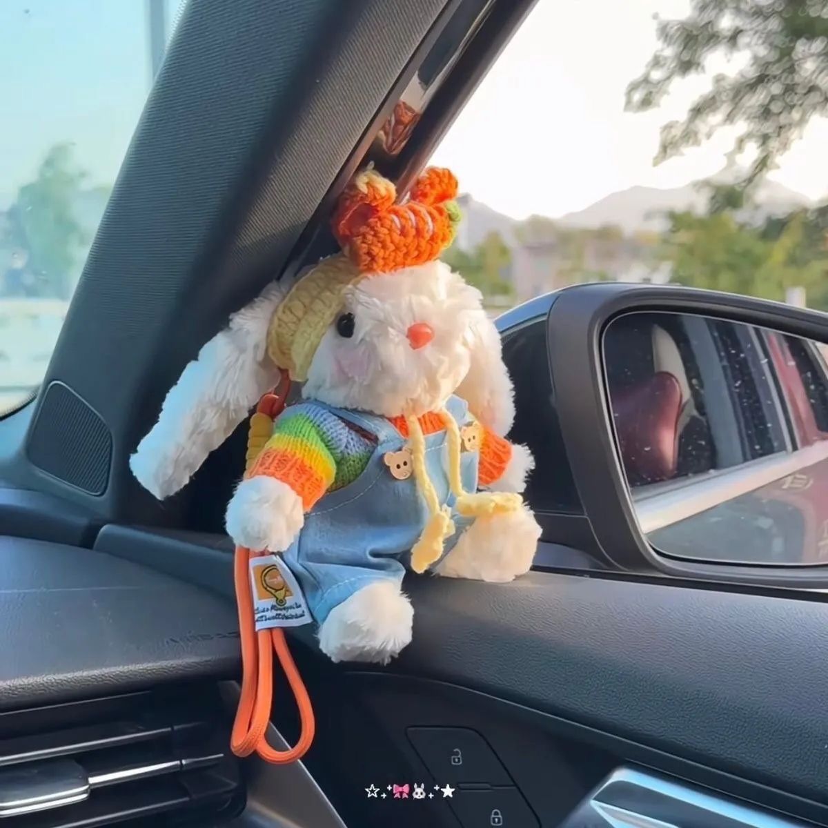 A white plush rabbit bag charm with long floppy ears, wearing a colorful striped sweater and blue overalls, topped with a crocheted orange hat, hanging from a car's rearview mirror as a cute and trendy car accessory.