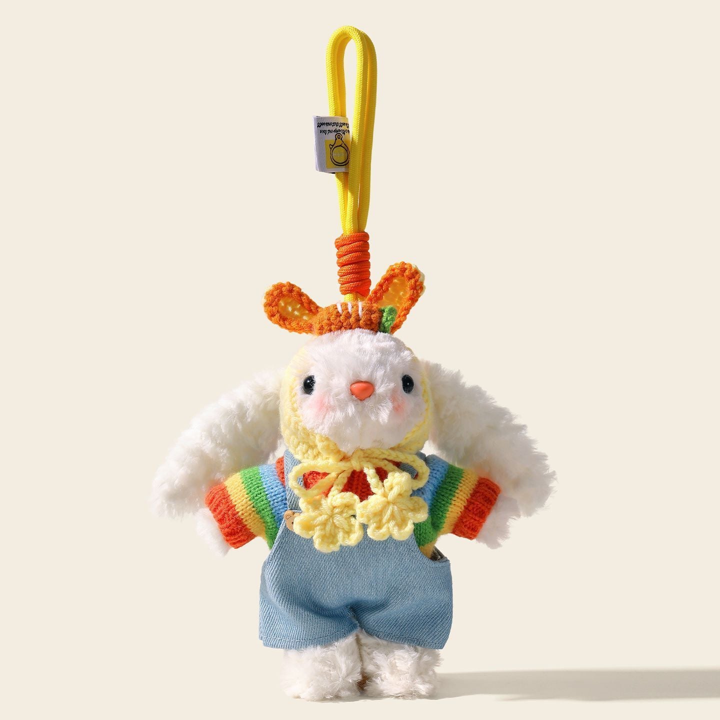 A white plush rabbit bag charm with long floppy ears, wearing a colorful striped sweater and blue overalls, topped with a crocheted orange hat. It features a yellow strap with a small tag, designed as a cute and trendy bag charm or car accessory.