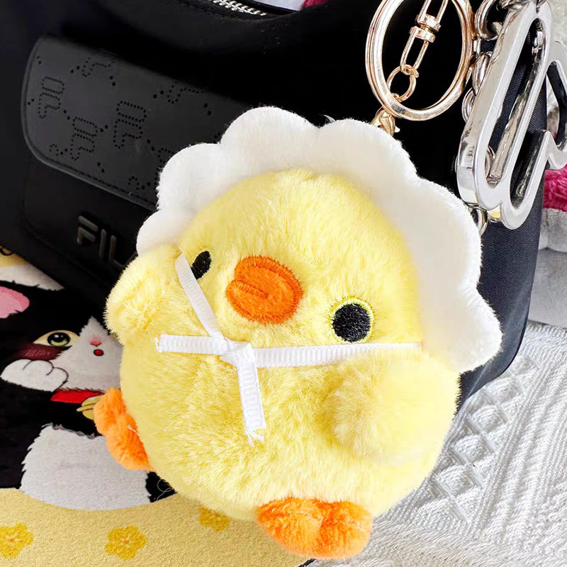 A yellow plush chick keychain in a white eggshell with a white ribbon bow, attached to a black bag with other keychains, displayed on a textured surface with a cartoon cat design nearby.