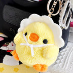 A yellow plush chick keychain in a white eggshell with a white ribbon bow, attached to a black bag with other keychains, displayed on a textured surface with a cartoon cat design nearby.