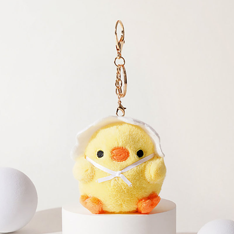 A yellow plush chick keychain emerging from a white eggshell, with black embroidered eyes, an orange beak, and orange feet, adorned with a white ribbon bow, attached to a gold-tone metal carabiner and keyring.