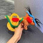 A hand holding two quirky plush keychains. The left one is a yellow creature wearing green underwear, and the right one is an orange horse-like figure with a blue headband and wild black hair. Both have large red lips and googly eyes.