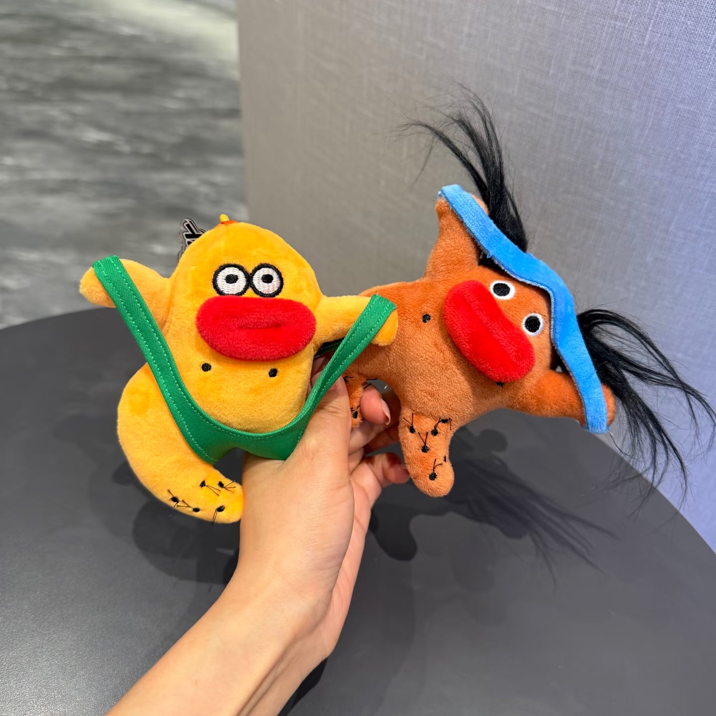 A hand holding two quirky plush keychains. The left one is a yellow creature wearing green underwear, and the right one is an orange horse-like figure with a blue headband and wild black hair. Both have large red lips and googly eyes.