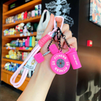 A hand holding a set of bright pink fitness-themed keychains, featuring a barbell plate, dumbbells, and a shaker bottle, with a blurred store shelf background.
