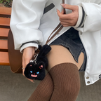 A person holding a brown leather bag with a fluffy black cat plush bag charm attached. The charm features bright blue eyes and a playful expression, adding a whimsical touch to the bag.