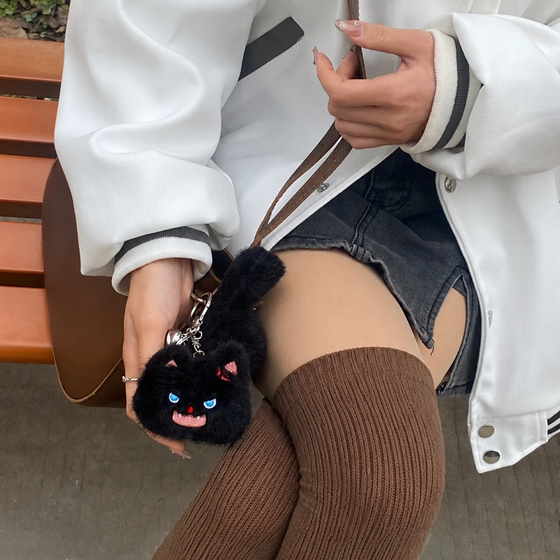 A person holding a brown leather bag with a fluffy black cat plush bag charm attached. The charm features bright blue eyes and a playful expression, adding a whimsical touch to the bag.