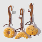 A set of three cute, food-themed plush keychains against a white background. From left to right: a yellow donut, a yellow croissant, and a yellow cookie with chocolate chips. Each has a smiling face and is attached to a brown braided rope with a metal clip and a small tag.