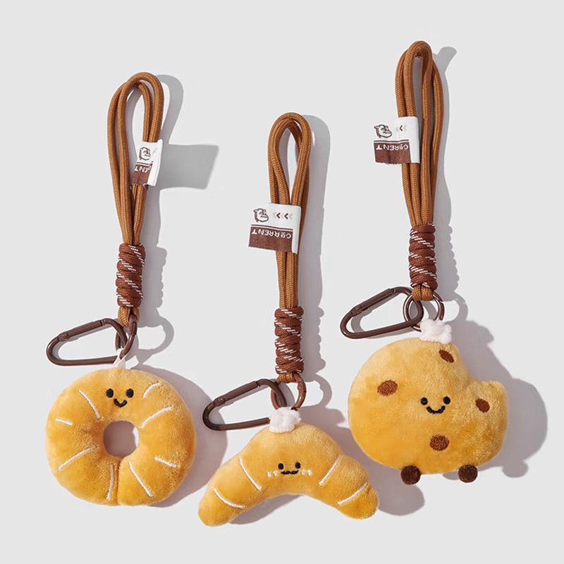 A set of three cute, food-themed plush keychains against a white background. From left to right: a yellow donut, a yellow croissant, and a yellow cookie with chocolate chips. Each has a smiling face and is attached to a brown braided rope with a metal clip and a small tag.