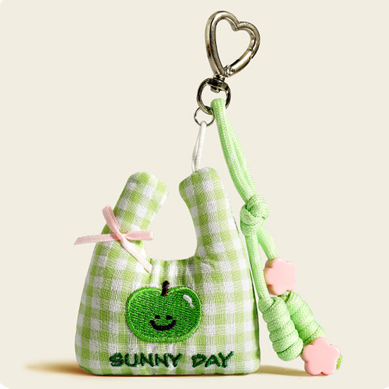 A green and white plaid fabric bag charm shaped like a mini tote, with an embroidered green apple and "SUNNY DAY" text, decorated with a pink bow and pink flower beads.