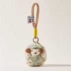 A front-facing view of a light green fluffy sheep plush keychain with brown spiral horns and a smiling face, attached to a beige cord with a yellow wrap and a small branded tag.