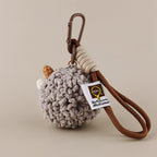 The back view of a grey crocheted sheep keychain showing its fluffy texture and a white brand tag attached to a brown rope strap and carabiner.