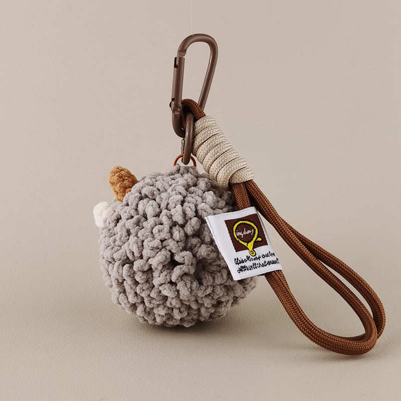The back view of a grey crocheted sheep keychain showing its fluffy texture and a white brand tag attached to a brown rope strap and carabiner.