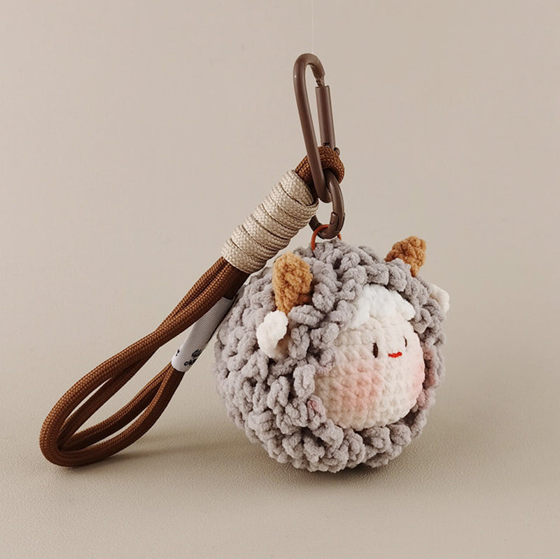 A grey crocheted sheep keychain with brown horns hanging from a brown strap against a neutral background.