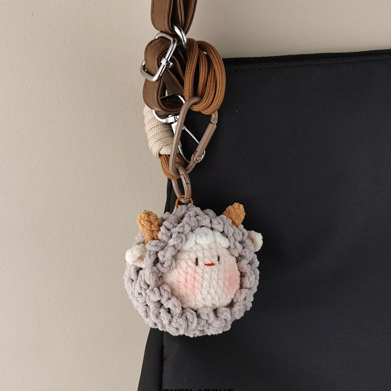 A grey crocheted sheep keychain with brown horns attached to a black bag with a brown strap and metal clips.