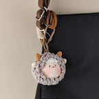 A grey crocheted sheep keychain with brown horns attached to a black bag with a brown strap and metal clips.