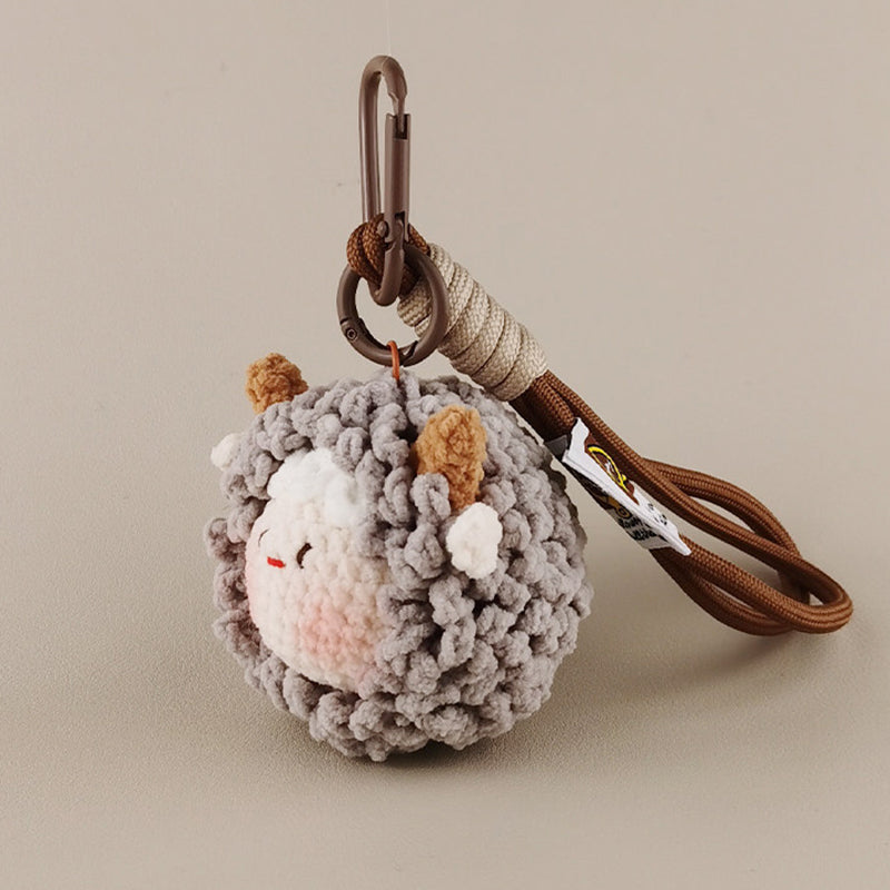 A side-angle shot of a grey crocheted sheep keychain with brown horns and white cheeks, hanging from a brown strap.