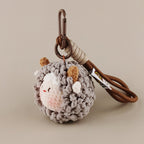 A side-angle shot of a grey crocheted sheep keychain with brown horns and white cheeks, hanging from a brown strap.