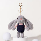 A grey plush rabbit keychain with long floppy ears, black embroidered eyes, and a pink nose, wearing a pink polka-dot shirt and a dark blue corduroy vest, attached to a gold-tone metal carabiner and keyring, displayed on a white pedestal.