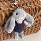 A grey plush rabbit keychain with long floppy ears, wearing a pink polka-dot shirt and a dark blue corduroy vest, attached to a gold-tone metal carabiner and hanging from a brown wicker bag.