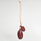 A hanging view of a handcrafted leather charm shaped like two coffee beans. The beans are dark brown with lighter stitching details and are suspended from a natural tan leather cord against a white background.