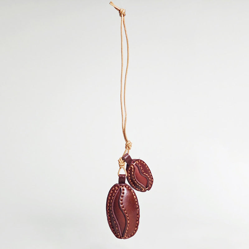 A hanging view of a handcrafted leather charm shaped like two coffee beans. The beans are dark brown with lighter stitching details and are suspended from a natural tan leather cord against a white background.