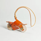 A close-up of a handmade leather motorcycle keychain in orange. It features brass rivets for wheels and is attached to a natural leather cord, displayed against a plain white background.