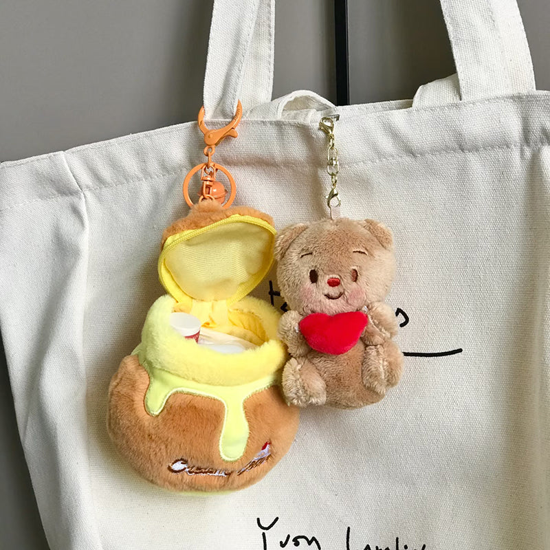 A person holding a white tote bag with two plush bear bag charms attached. One is a brown bear holding a red heart, and the other is a brown bear nestled inside a honey jar-shaped container. Both are attached to orange lobster clasps and keyrings.