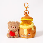 A detachable plush teddy bear bag charm in brown, holding a red heart, shown separately from its honey jar-shaped container. The bear can be removed from the jar, which has a zipper closure and is attached to an orange lobster clasp and keyring.