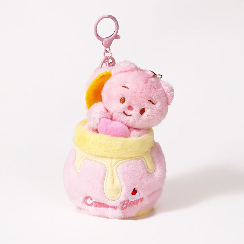 A plush teddy bear bag charm in pink, holding a pink heart, nestled inside a honey jar-shaped container with a yellow rim and dripping honey details. The jar has a zipper closure and is attached to a pink lobster clasp and keyring, designed as a cute and sweet accessory for bags or keychains.