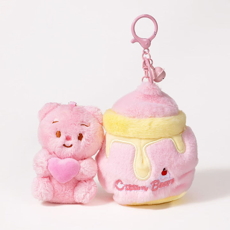 A detachable plush teddy bear bag charm in pink, holding a pink heart, shown separately from its honey jar-shaped container. The bear can be removed from the jar, which has a zipper closure and is attached to a pink lobster clasp and keyring.