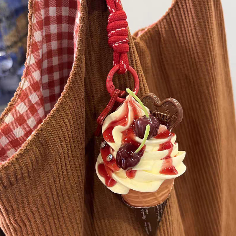 A cute, yellow plush ice cream cone keychain with white whipped cream, red strawberry sauce, and two cherries on top. It is attached to a red braided rope with a metal ring and a small white tag featuring a logo.