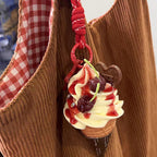A cute, yellow plush ice cream cone keychain with white whipped cream, red strawberry sauce, and two cherries on top. It is attached to a red braided rope with a metal ring and a small white tag featuring a logo.